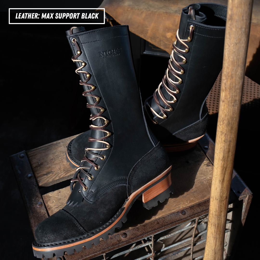 A pair of black leather lace-up boots with gold-tone eyelets and lug soles, displayed on a rustic wooden crate with a wooden pole in the foreground. A text overlay in the top-left corner reads 'LEATHER: MAX SUPPORT BLACK'.