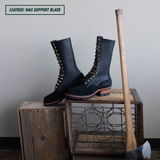 A pair of tall black leather lace-up boots displayed on rustic wooden crates, with a wooden-handled tool leaning against the crate. A text label in the top-left corner reads 'LEATHER: MAX SUPPORT BLACK'.
