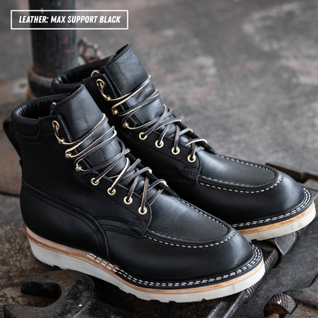 A pair of black leather lace-up ankle boots with white stitching and white soles, resting on a dark textured metal surface. A text overlay in the top-left corner reads 'LEATHER: MAX SUPPORT BLACK'.