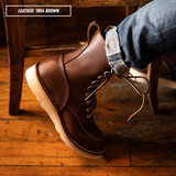 A brown leather lace-up work boot worn on a foot with cuffed blue jeans, photographed in a rustic indoor setting with wooden furniture and floorboards. A text label in the top left reads 'LEATHER: 1964 BROWN'.