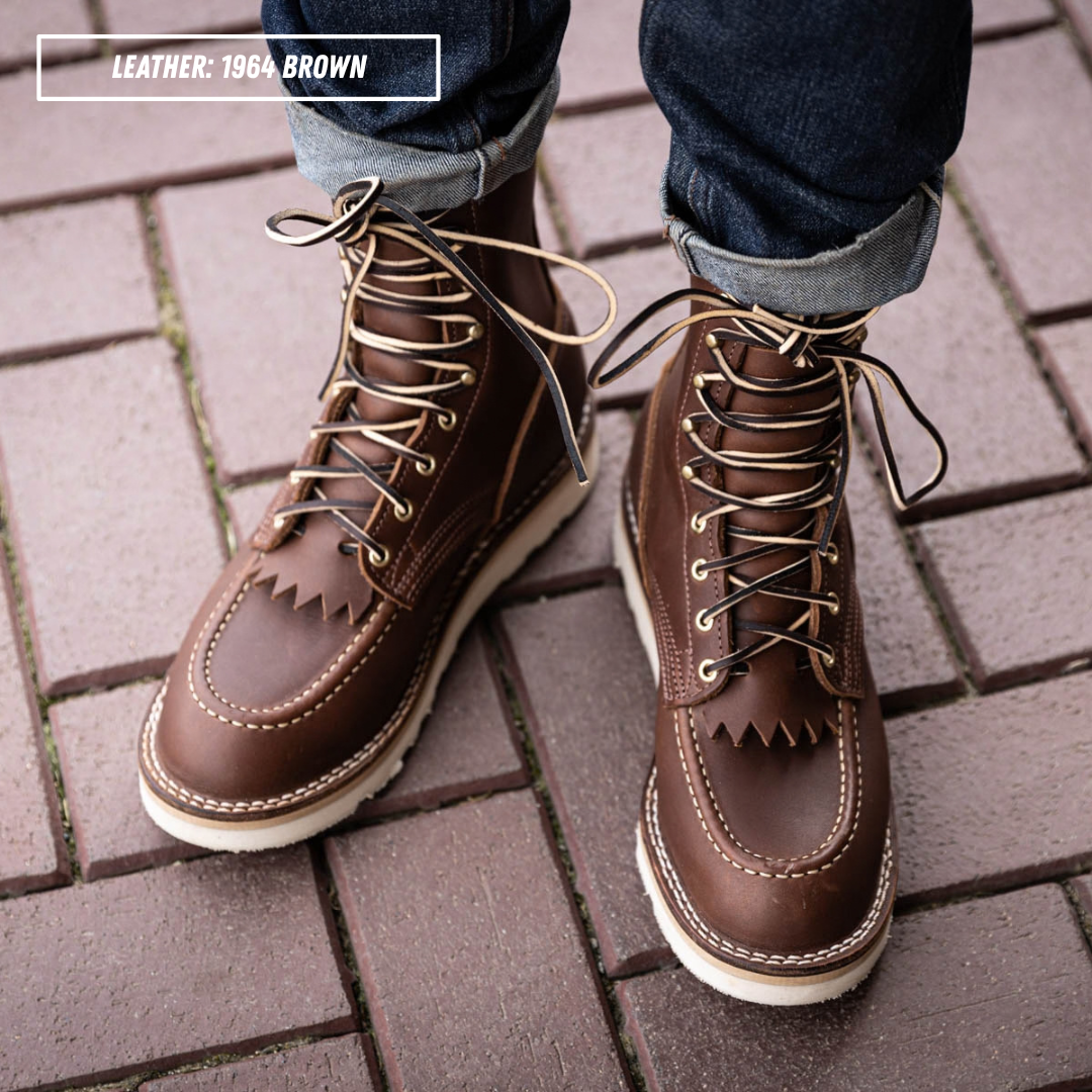 A pair of men's brown leather lace-up boots with fringe detailing on the vamp, shown worn with cuffed blue jeans on a brick pavement. A text overlay in the top left reads 'LEATHER: 1964-BROWN'.
