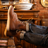A pair of brown leather lace-up boots worn with dark blue jeans, shown with one boot standing and the other resting on a wooden surface in a rustic indoor setting. A text overlay in the top left reads 'LEATHER: 1964 BROWN'.