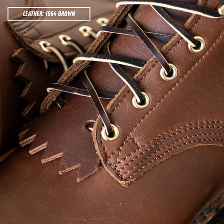 Close-up of a brown leather lace-up boot with white laces and brass eyelets, showing the textured leather and stitching. A text label in the top left reads 'LEATHER: 1964 BROWN'.
