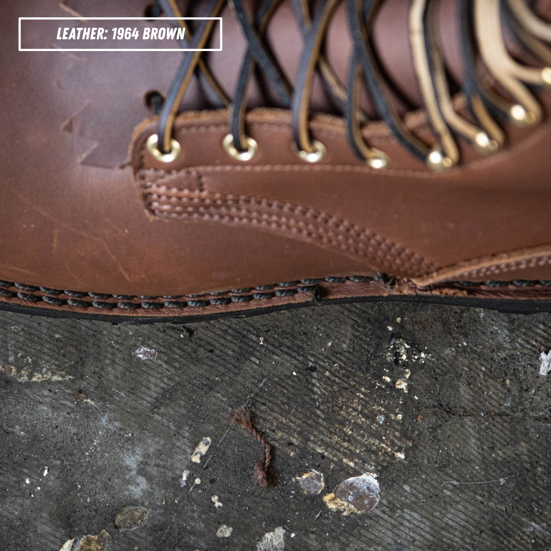 Close-up of a brown leather lace-up boot corner with metal eyelets and a thick stitched welt, shown resting on a dark textured surface. A label in the top left reads 'LEATHER: 1964 BROWN'.