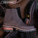 A dark brown leather Chelsea-style ankle boot with a chunky stacked heel, black lug sole, and a light tan midsole section laying on a pile of other Chelsea boot. Top left reads "LEATHER: 1964 MAHOGANY."