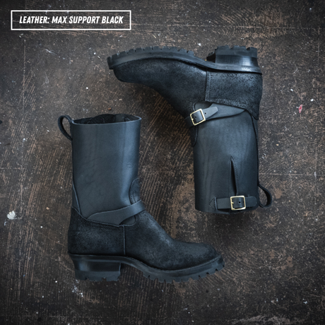 A pair of black leather mid-calf boots with buckle straps, displayed on textured concrete. Boots are lying on their sides, showing the side profile and buckle details.