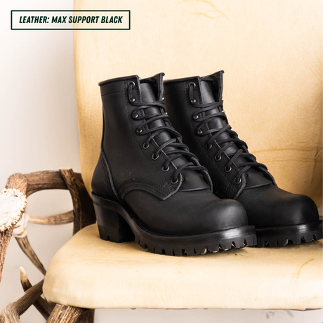 A pair of black leather lace-up ankle boots with chunky lug soles, displayed on a beige upholstered chair with rustic wooden branches. A text overlay in the top-left corner reads 'LEATHER: MAX SUPPORT BLACK'.