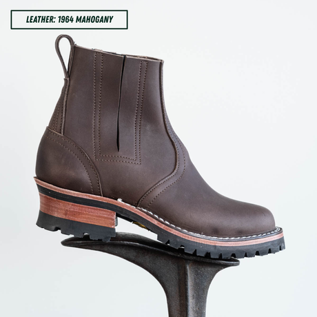 A dark brown leather ankle boot with a chunky block heel, displayed on a black metal stand against a light gray background. A text label in the top-left corner reads 'LEATHER: 1964 MAHOGANY'.