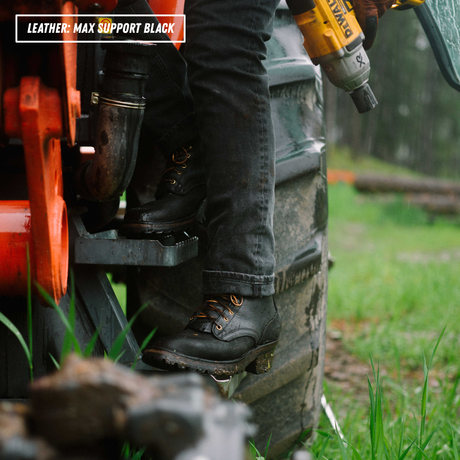 A close-up outdoor shot of a person wearing black leather work boots with orange laces, standing on the steps of a tractor next to a large tire. A yellow DeWalt cordless drill is held in the person's hand, and a text overlay in the top-left corner reads 'LEATHER - MAX SUPPORT BLACK'.