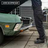A person wearing black leather work pants stands beside an open green car hood in a driveway. The text overlay at the top left reads 'LEATHER: MAX SUPPORT BLACK'.