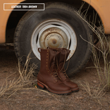 A pair of 1964-style brown leather lace-up work boots placed in front of a vintage vehicle wheel on dry grass and dirt ground. A text label in the top-left reads 'LEATHER: 1964 BROWN'.