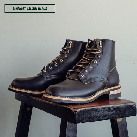 A pair of dark brown leather lace-up boots with tan laces and gold-toned eyelets, displayed on a rustic wooden stool against a light gray wall. A text label in the top left reads 'LEATHER: GALLUN BLACK'.