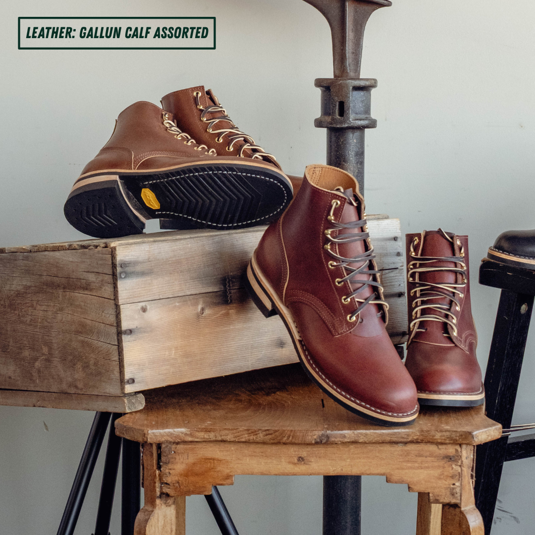 Two pairs of brown leather lace-up work boots displayed on rustic wooden crates and stools, with one boot showing its black treaded sole. Text in the top-left corner reads 'LEATHER: GALLUN CALF ASSORTED'.