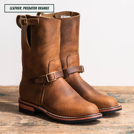 A pair of mid-calf leather boots in a warm brown shade, featuring a buckle strap across the instep and pull tabs at the top, displayed on a rustic wooden surface against a light background. A text overlay in the top left corner reads 'LEATHER: PREDATOR ORANGE'.