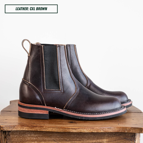 A pair of dark brown leather Chelsea boots with black elastic side panels, displayed on a rustic wooden block against a white background. A text label in the top left reads 'LEATHER: CXL BROWN'.