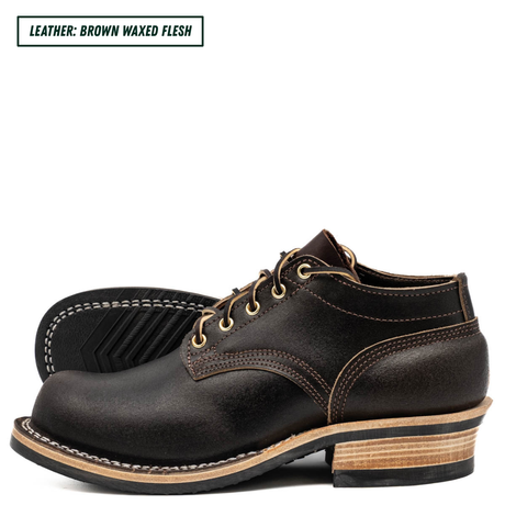 A pair of men's lace-up work boots in dark brown waxed leather, shown from a side profile view with one boot standing upright and the other lying flat to display the sole. The boots feature contrast stitching, a stacked wood-look heel, and a black rubber outsole.