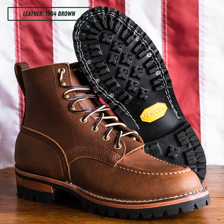 A pair of brown leather lace-up work boots shown on a wooden surface with an American flag background. One boot is standing upright, and the other is turned to show the black treaded sole with a yellow Vibram logo.
