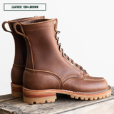 A pair of brown leather lace-up boots with metal eyelets and thick lug soles, displayed on a rustic wooden surface against a white background. A text label in the top left reads 'LEATHER: 1964 BROWN'.