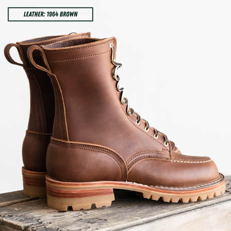 A pair of brown leather lace-up boots with metal eyelets and thick lug soles, displayed on a rustic wooden surface against a white background. A text label in the top left reads 'LEATHER: 1964 BROWN'.