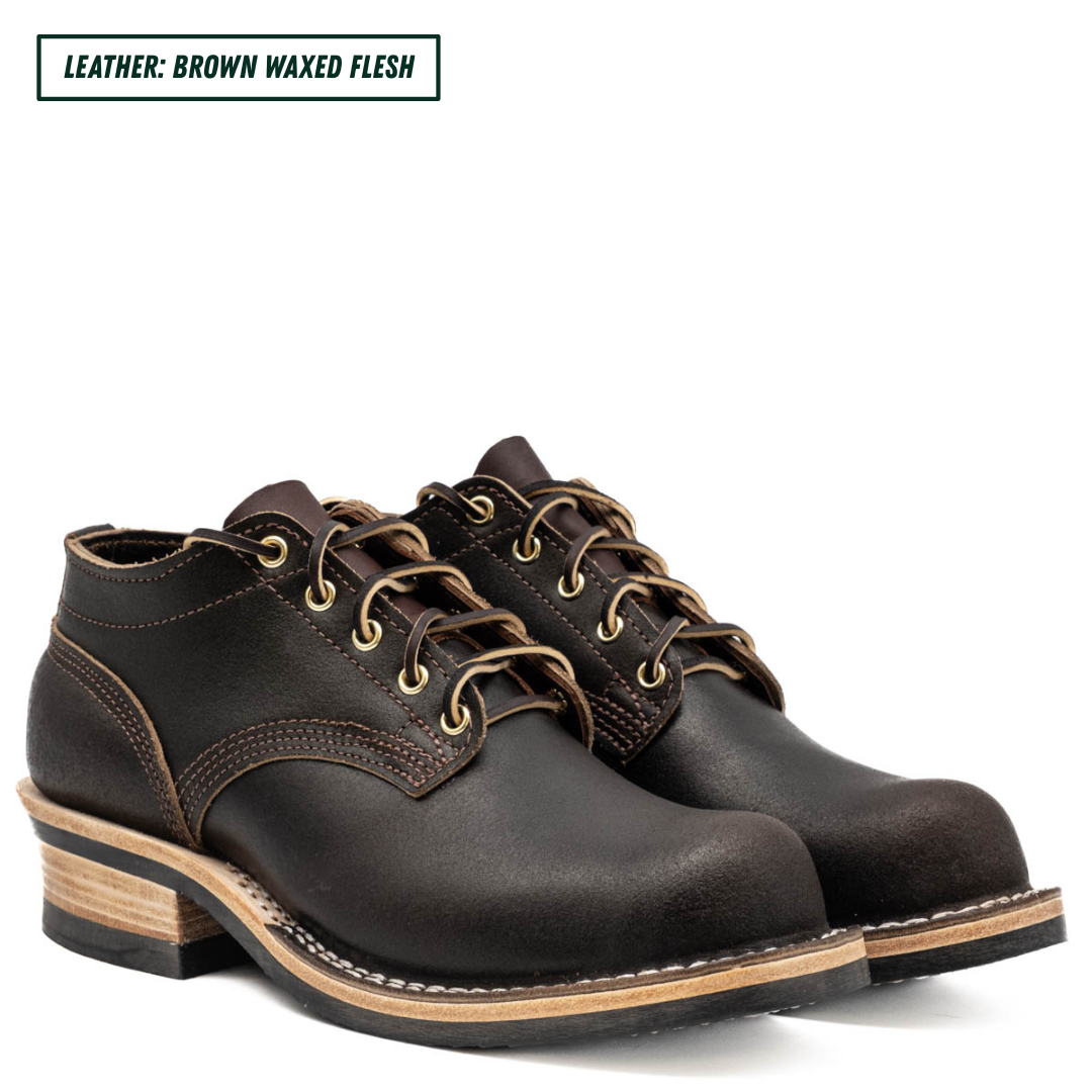 A pair of dark brown leather lace-up work boots with stacked wood-look heels, shown on a white background with a text label in the top-left corner reading 'LEATHER: BROWN WAXED FLESH'.