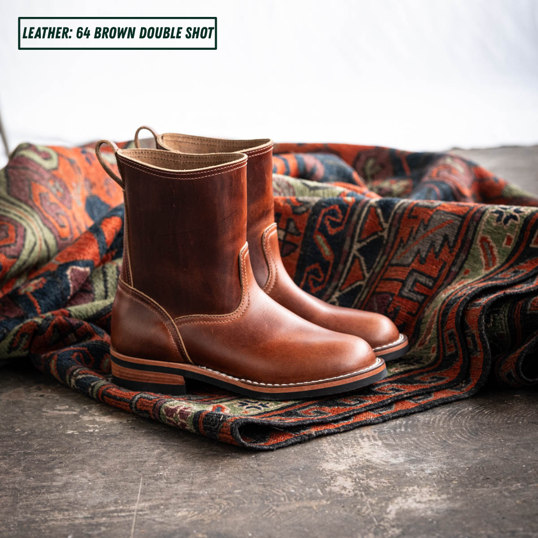 A pair of brown leather ankle boots displayed on a patterned rug over a dark wooden floor, with a text overlay in the top left reading 'LEATHER: 64 BROWN DOUBLE SHOT'.