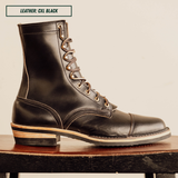 A single black leather lace-up ankle boot shown in side profile, resting on a wooden surface against a plain light beige background. A small label at the top reads 'LEATHER: CXL BLACK'.