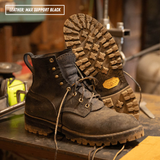 A pair of rugged lace-up work boots in dark brown leather, with one boot standing upright and the other lying on its side to show the sole. The boots are placed on a workbench in a workshop setting.