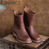 A pair of mid-calf brown leather boots with white contrast stitching, displayed on a rustic wooden crate against a textured brown backdrop. A text overlay in the top left reads 'LEATHER: 1964 BROWN'.