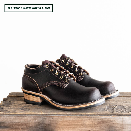 A pair of dark brown leather lace-up shoes with stacked wood-look heels, displayed on a rustic wooden surface against a white background. A text label in the top-left corner reads 'LEATHER: BROWN WAXED FLESH'.