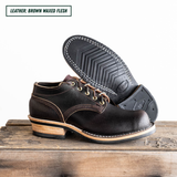 A pair of dark brown waxed leather lace-up boots, with one boot standing upright and the other tilted to show the treaded outsole. They are displayed on a rustic wooden surface against a white background, with a text label in the top-left corner reading 'LEATHER: BROWN WAXED FLESH'.