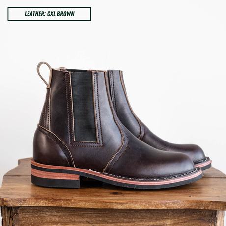 A pair of dark brown leather Chelsea boots with black elastic side panels, displayed on a rustic wooden block against a white background. A text overlay in the top left reads 'LEATHER: CXL BROWN'.