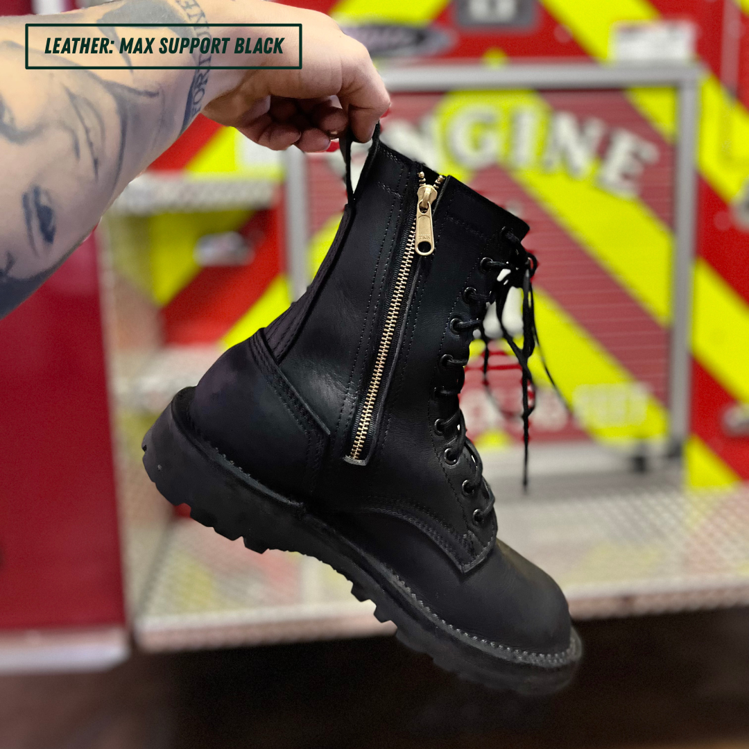 A hand holds up a black leather lace-up boot with a prominent gold zipper on the side. The background shows a red and yellow striped fire truck interior, and a text overlay in the top left reads 'LEATHER: MAX SUPPORT BLACK'.