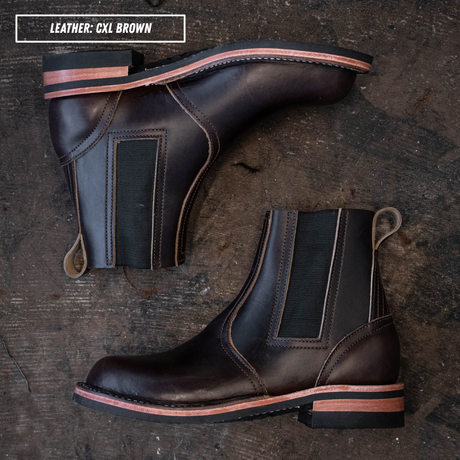 Two dark brown leather Chelsea boots arranged on a rustic dark wood surface, with a text label in the top-left corner reading 'LEATHER: CXL BROWN'. The boots have elastic side panels, pull tabs, and a low stacked heel with a contrasting tan edge.