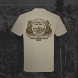 A light beige crewneck t-shirt shown from the back, featuring a large brown and gold graphic print with the text 'Stronger Together Wildfire Relief Effort' and 'Spokane County, Washington'. The shirt is displayed against a dark, moody forest background.