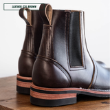 A pair of dark brown Chelsea boots made of cxl brown leather, shown from a rear three-quarter angle on a wooden surface against a white background. A text overlay in the top left corner reads 'LEATHER: CXL BROWN'.