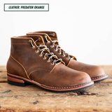 A pair of rugged brown leather lace-up boots with a distressed finish, displayed on a rustic wooden surface against a white background. A text label in the top left reads 'LEATHER: PREDATOR ORANGE'.