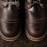 Close-up of a pair of dark brown leather lace-up shoes with white contrast stitching and a textured finish. A text label in the top left reads 'LEATHER: BROWN WAXED FLESH'.