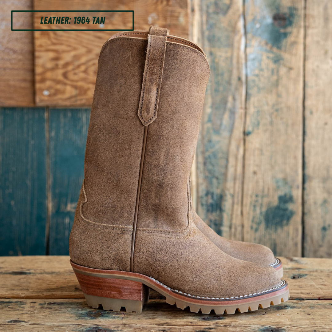 A pair of tan suede western-style boots shown in side profile on a wooden table with a wood pallet background. A small label in the top left reads 'LEATHER: 1964 TAN'.