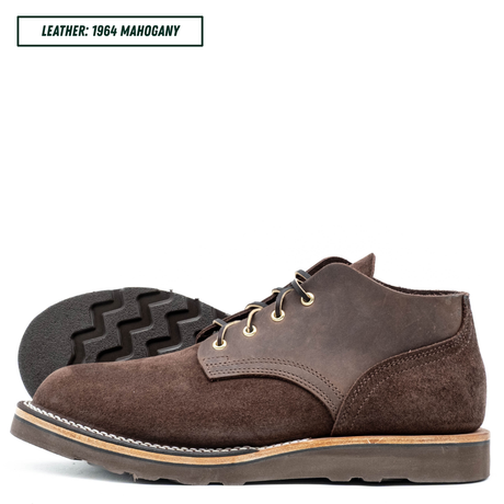 A pair of dark brown suede and leather lace-up boots shown in profile, with one boot standing upright and the other lying on its side to display the treaded outsole. A text label in the top left reads 'LEATHER: 1964 MAGHOGANY'.