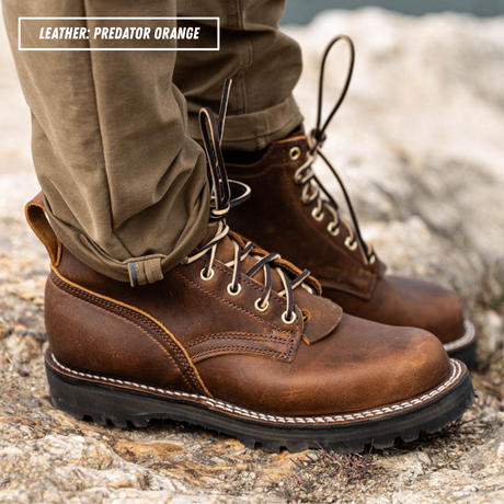 A close-up of a person wearing brown leather lace-up boots with cuffed khaki pants, standing on a rocky outdoor surface. A text label in the top left reads 'LEATHER - PREDATOR ORANGE'.