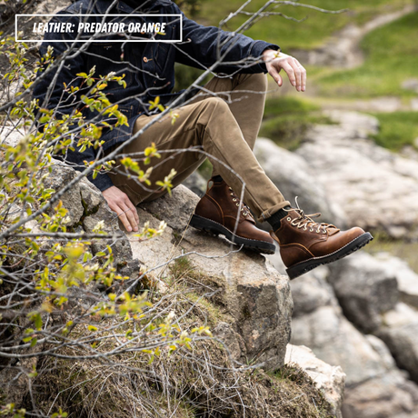 A person sitting on a rocky outdoor ledge wearing brown lace-up boots, tan pants, and a dark jacket, with a text overlay reading 'LEATHER: PREDATOR ORANGE'.