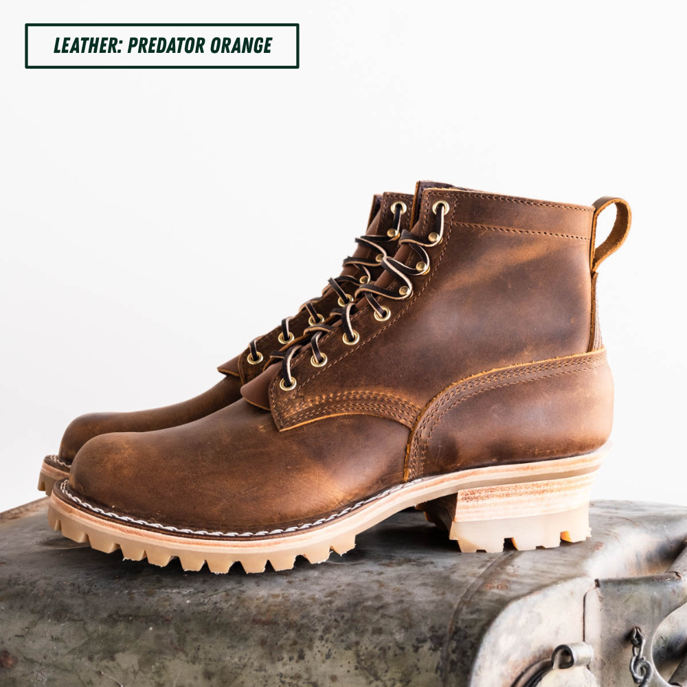 A pair of rugged lace-up ankle boots in distressed brown leather, displayed on a vintage metal trunk against a white background. A text label in the top left reads 'LEATHER: PREDATOR ORANGE'.