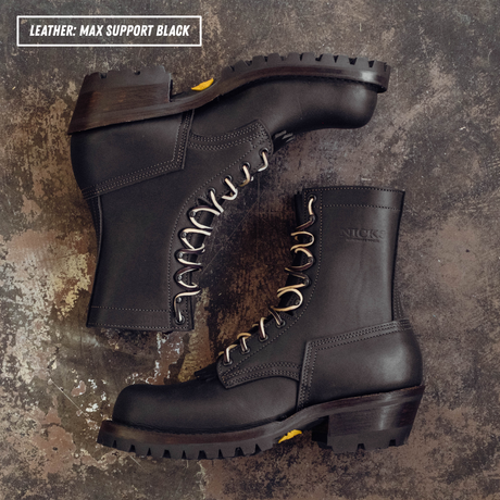 Two black leather work boots arranged on a textured dark surface. One boot is positioned upright and the other is angled on top, showing the side, toe, and laces.