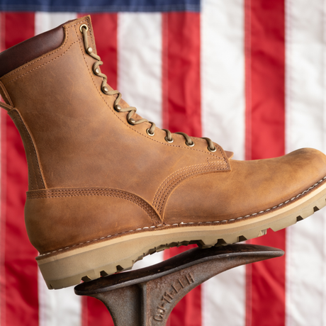 A tan leather lace-up work boot with a thick beige sole, shown in profile against a blurred American flag background, resting on a dark metal display stand.
