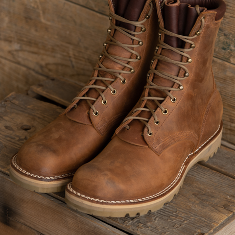 A pair of rugged brown leather lace-up boots with beige soles and metal eyelets, displayed on rustic wooden planks.