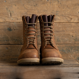 A pair of brown leather lace-up work boots with tan laces and light beige soles, displayed on a rustic wooden surface against a wooden plank background.