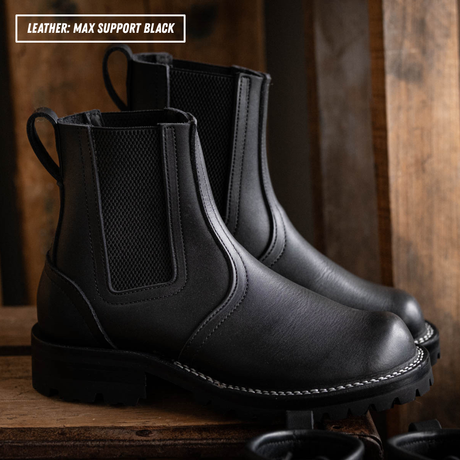 Blackout Chelsea boots sitting on wooden crate in front of pallet wall. Boots are shown in side profile with one propped up slightly. Top left reads 'LEATHER: MAX SUPPORT BlACK'.