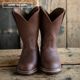 A pair of brown leather cowboy boots standing upright on a rustic wooden surface against a weathered wood plank background. A text overlay in the top left reads 'LEATHER: 1964 BROWN'.