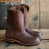 A pair of brown leather cowboy boots displayed on a rustic wooden surface with a wooden plank background. A text overlay in the top left corner reads 'LEATHER: 1964 BROWN'.