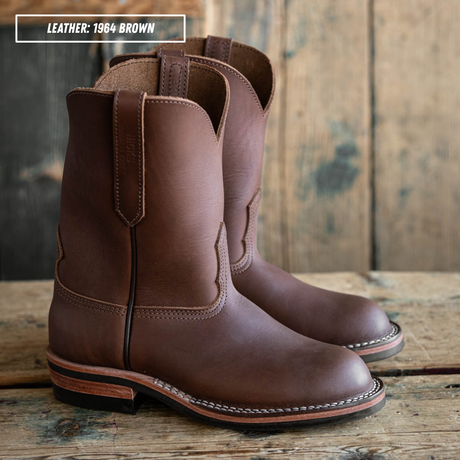 A pair of brown leather cowboy boots displayed on a rustic wooden surface with a wooden plank background. A text overlay in the top left corner reads 'LEATHER: 1964 BROWN'.